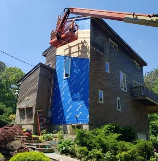 Two-story home mid-rebuild with blue house wrap and exposed wall prep on the side elevation.