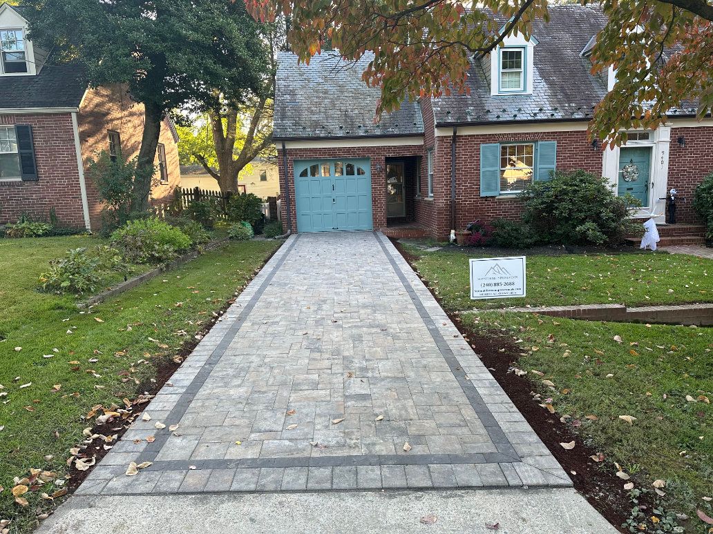 Brick paver walkway bordered by landscaped planting beds leading toward a home at dusk.
