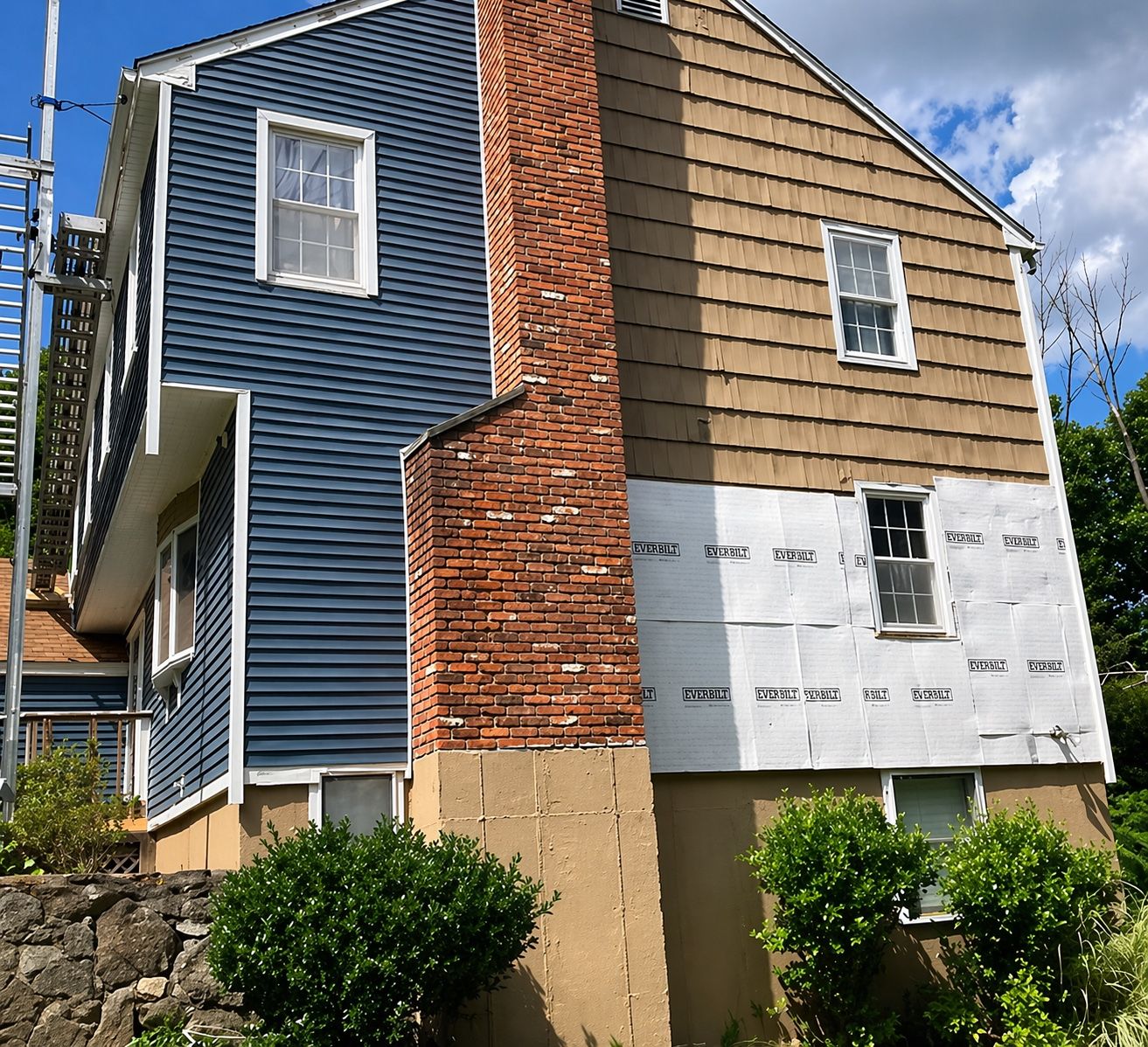 Exterior chimney rebuild in progress — exposed historic red brickwork, stucco foundation repair below, and Everbilt house wrap on the adjoining gable.
