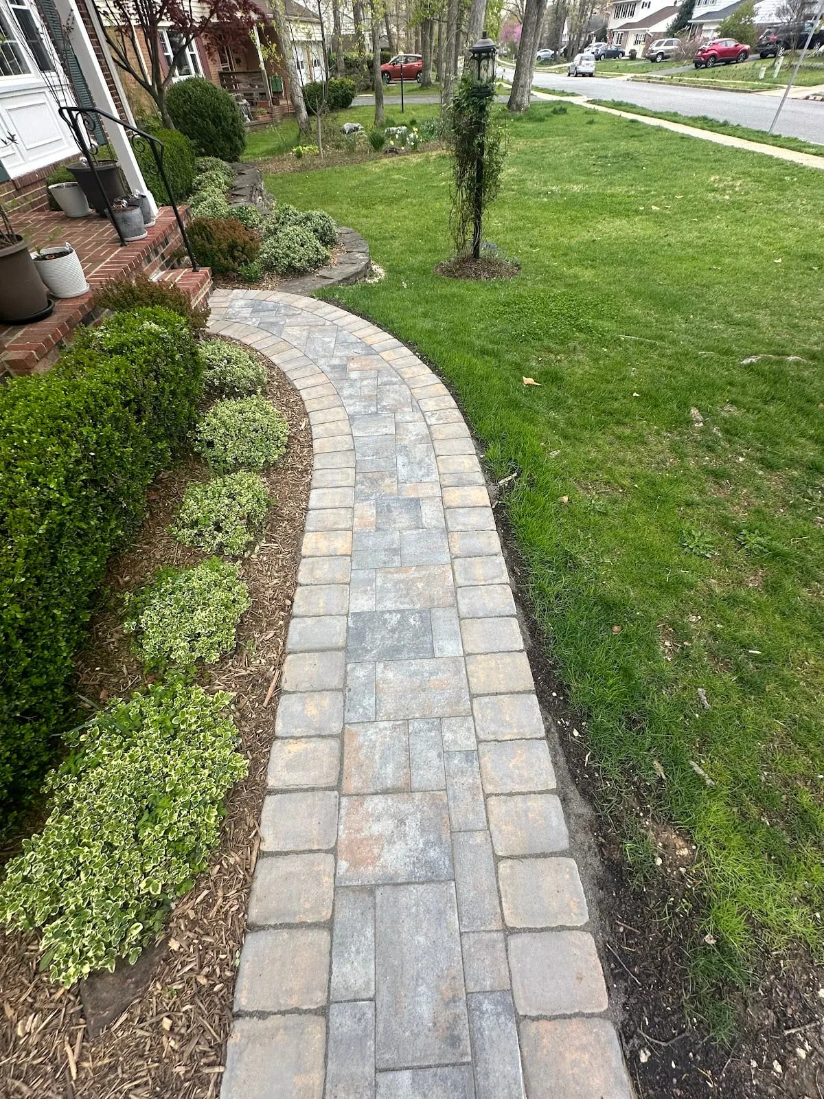 Curving multi-tone gray paver walkway with brick soldier border winding through lush garden beds along front of home