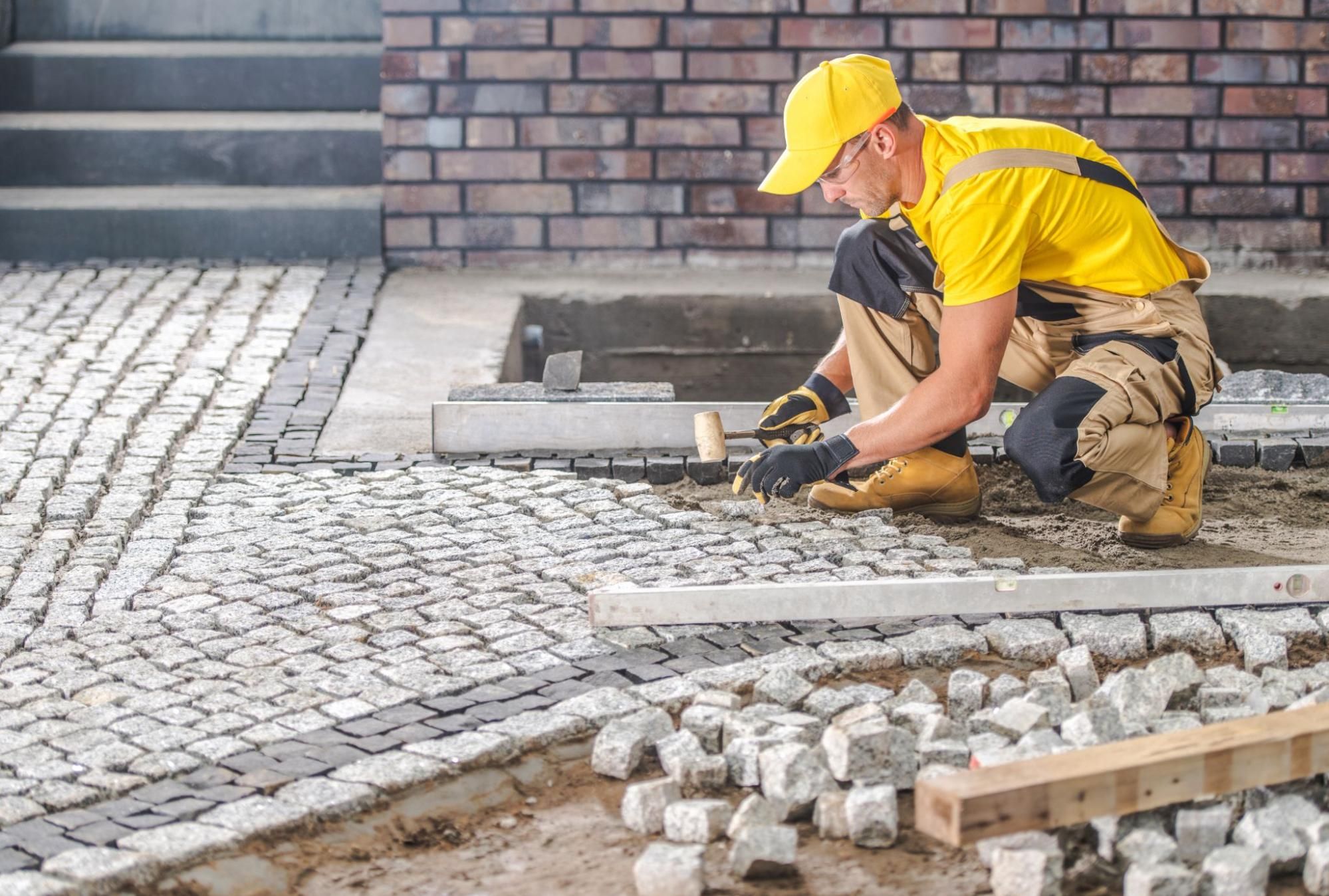 Stonemason kneeling on a sand base setting hand-cut cobblestone pavers along a garden edge.