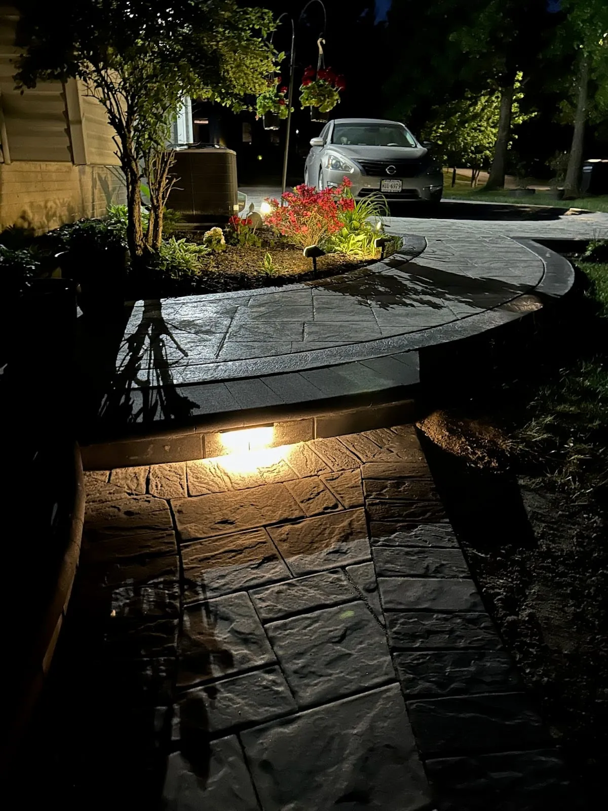 Curved paver walkway at night around a planted island with hanging-basket lamppost